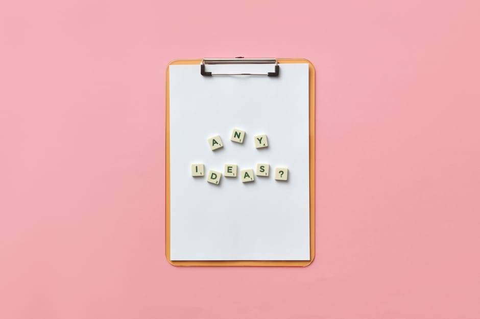 Creative conceptual image of a clipboard with letters spelling 'Any Ideas?' using Scrabble tiles on a pink background.